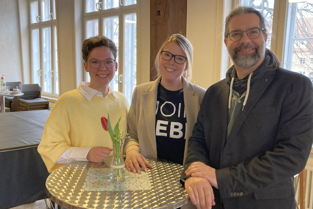 Zwei Frauen und ein Mann stehen an einem Bistrotisch und lächeln in die Kamera.
